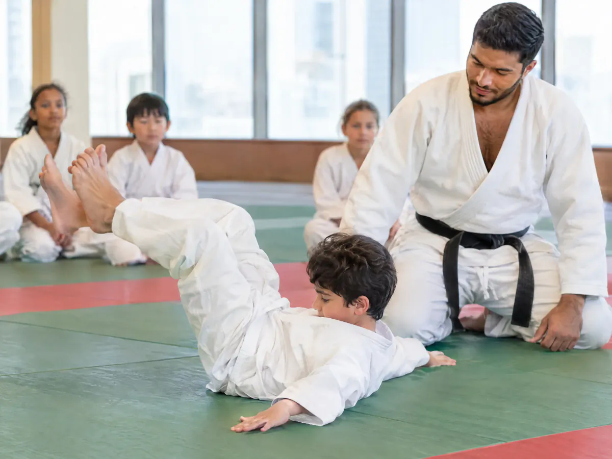 A boy in a white judo uniform rolls backwards onto the mat with his chin tucked and his arms out, as a coach kneels beside him, watching.