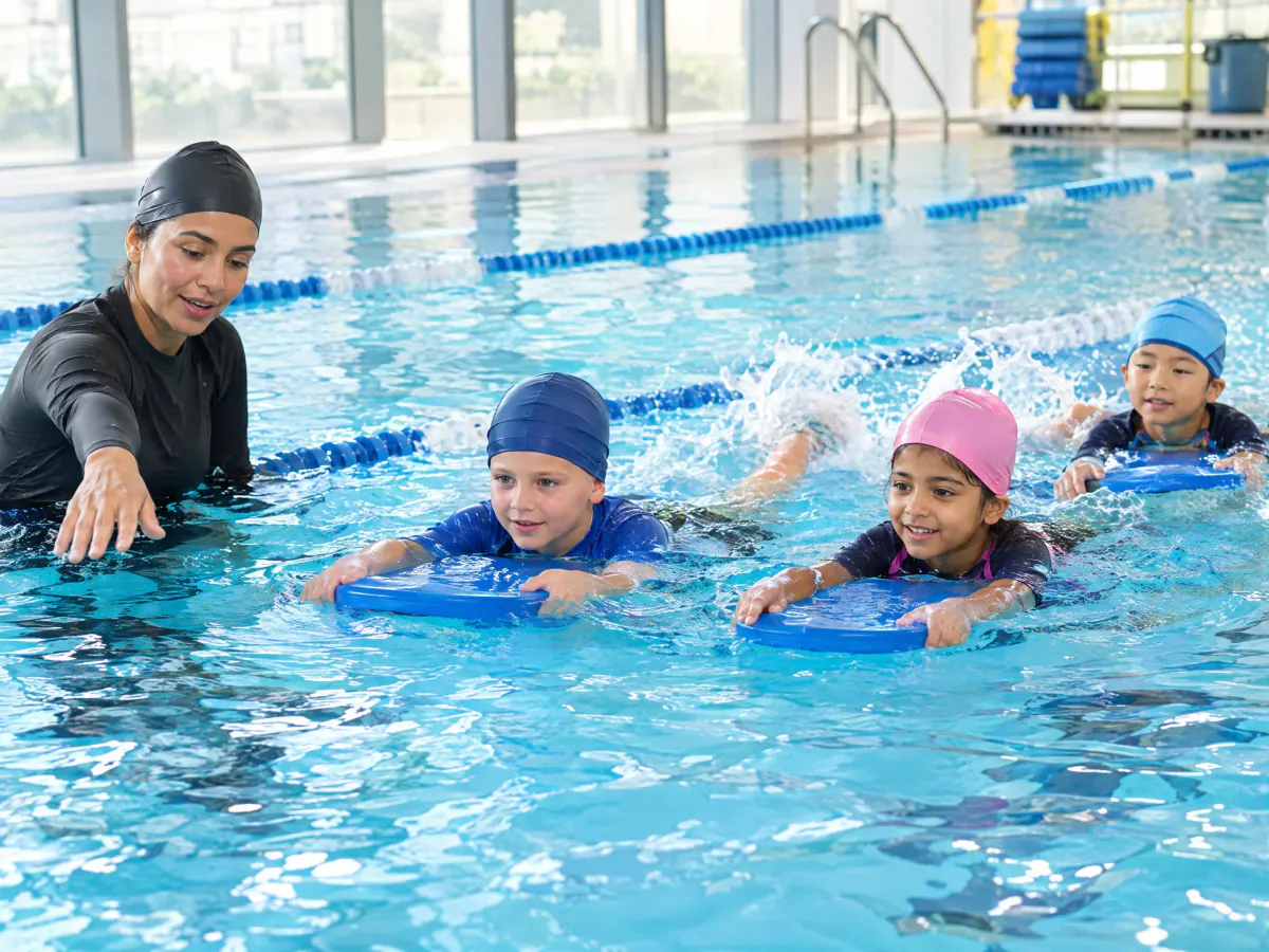 An instructor in an indoor learner pool stays within arm's reach of three children kicking along the lane with foam kickboards.