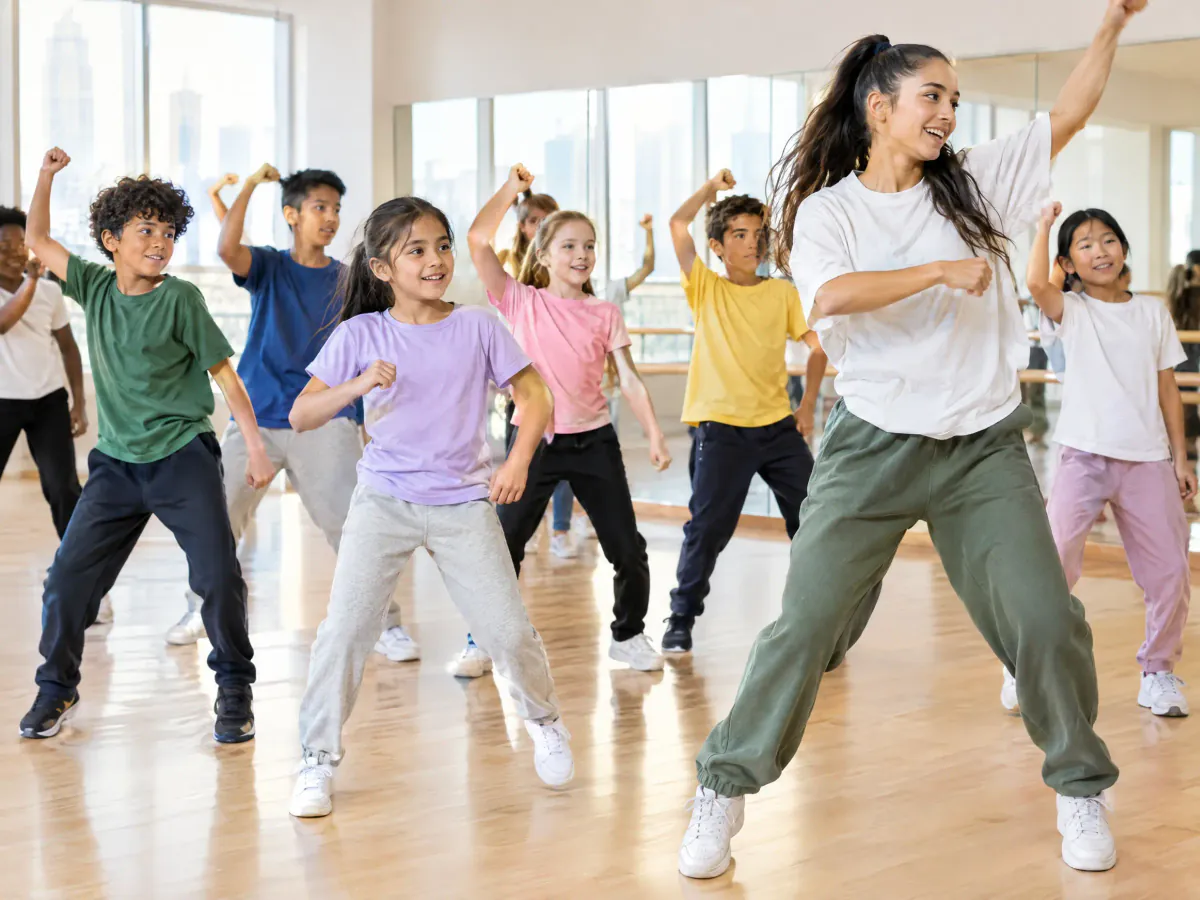 Children in T-shirts and joggers learn a street-dance routine in a bright studio, arms raised, following a teacher at the front.