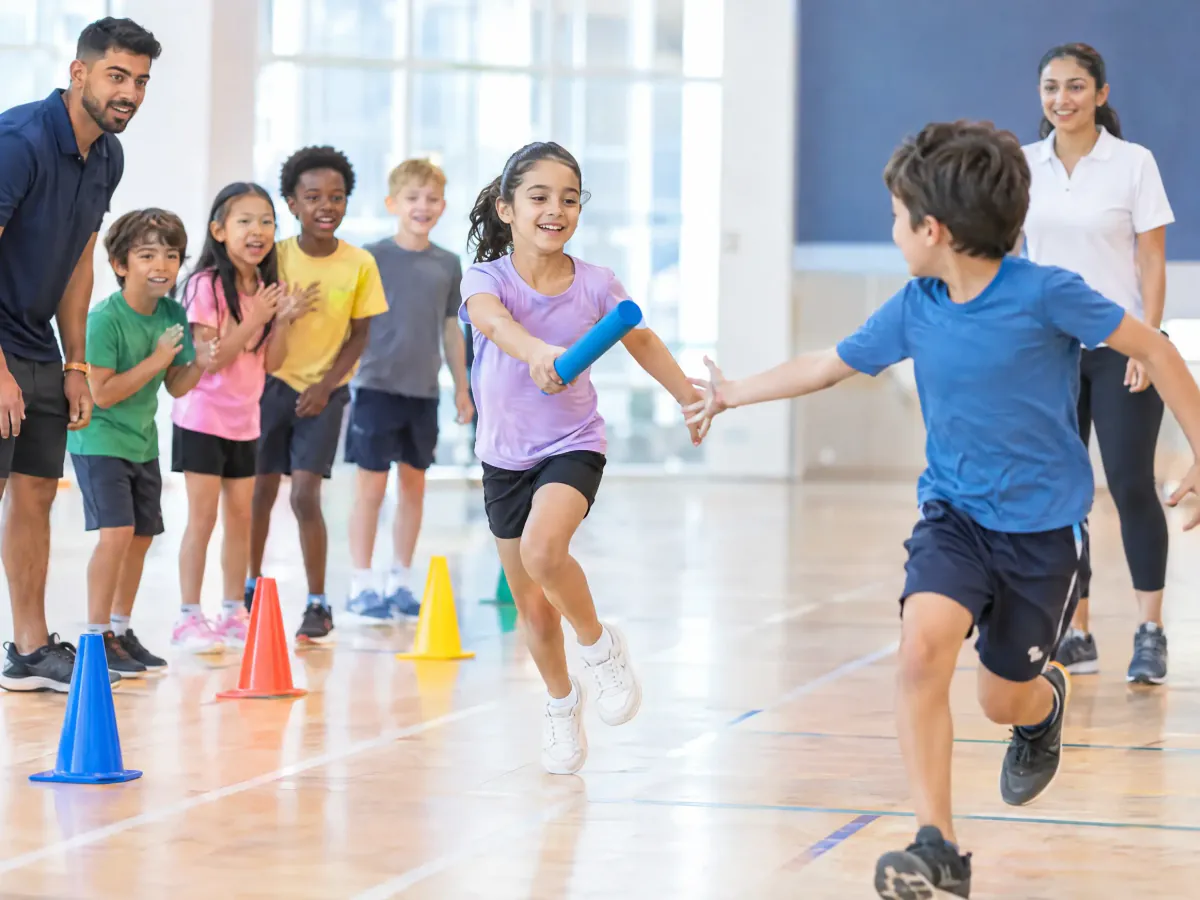 Children at a holiday camp in a sports hall run a relay, one passing a foam baton to the next while teammates cheer behind a line of cones.