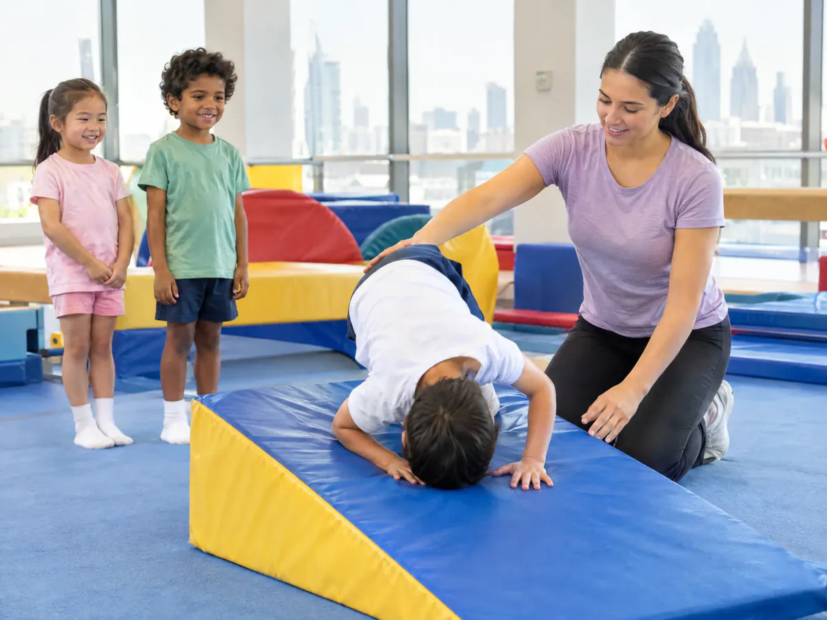 A young boy does a forward roll down a soft wedge mat while a coach kneels beside him with a hand on his back. Two children wait their turn.