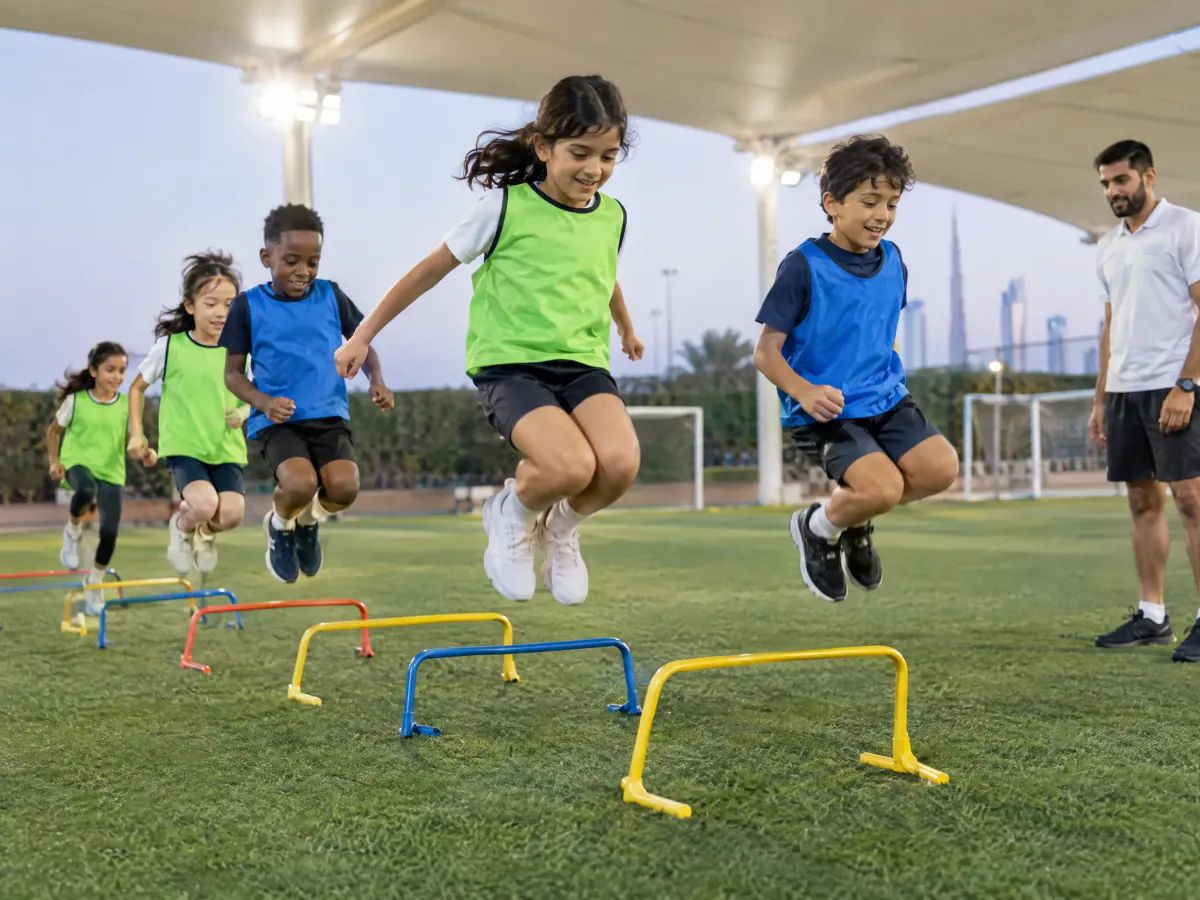 Boys and girls in training bibs jump over low coloured hurdles on an artificial pitch under a shade canopy, with a coach watching.