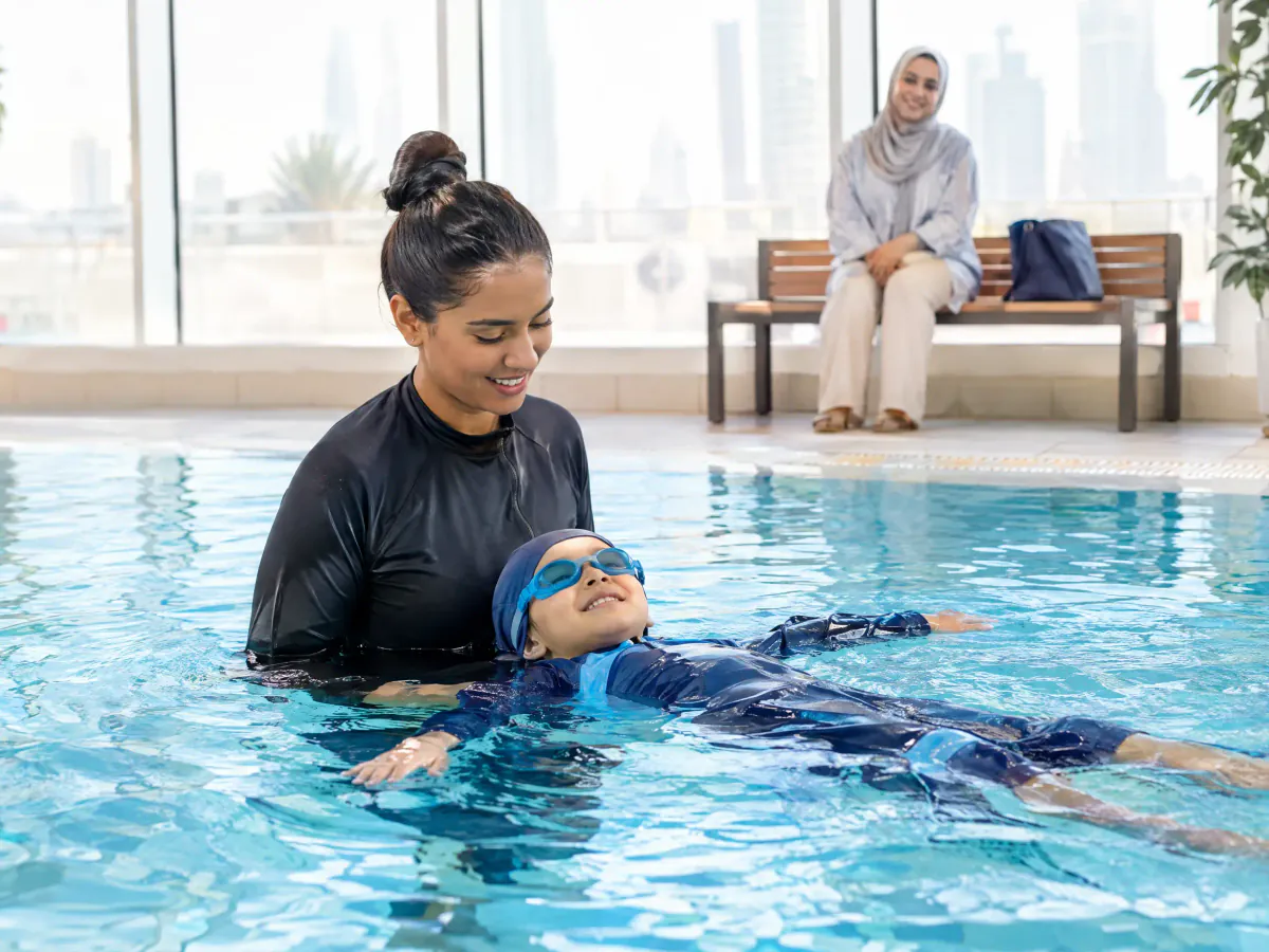 A swimming instructor in an indoor pool supports a young child floating on their back, while a parent watches from a bench at the poolside.