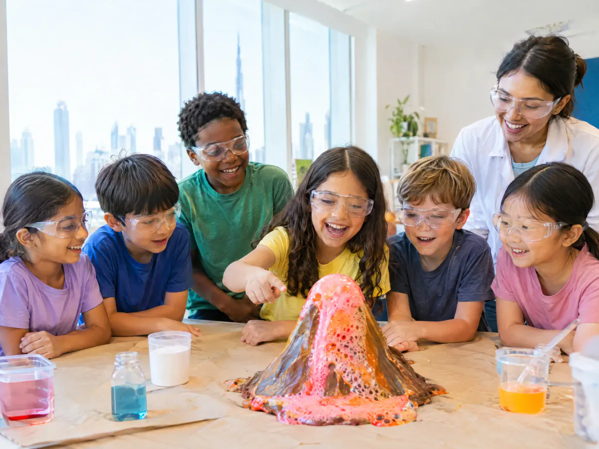 Children in safety goggles at a holiday science camp watch a colourful home-made volcano bubble over on a paper-covered table, with a camp leader beside them.