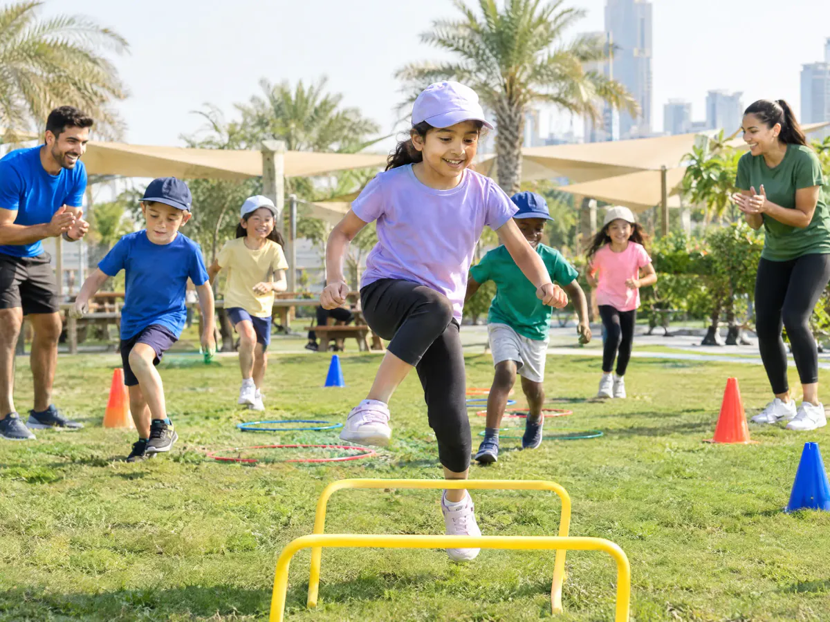 Children at a holiday camp in a green park run through an obstacle course of hoops, cones and low hurdles in winter sunshine, while two camp leaders cheer them on.