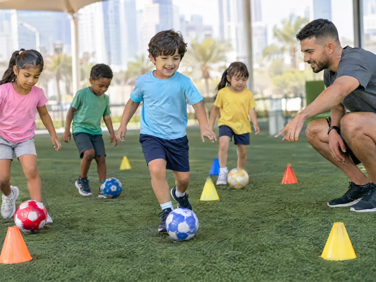 Four-year-olds dribble small footballs around coloured cones on an artificial pitch, with a coach crouching at their level and pointing the way.